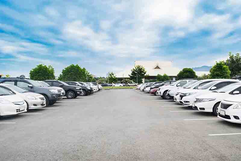 Car parking in large asphalt parking lot with trees, white cloud and blue sky background in front of hall building. Outdoor parking lot with fresh ozone and green environment concept